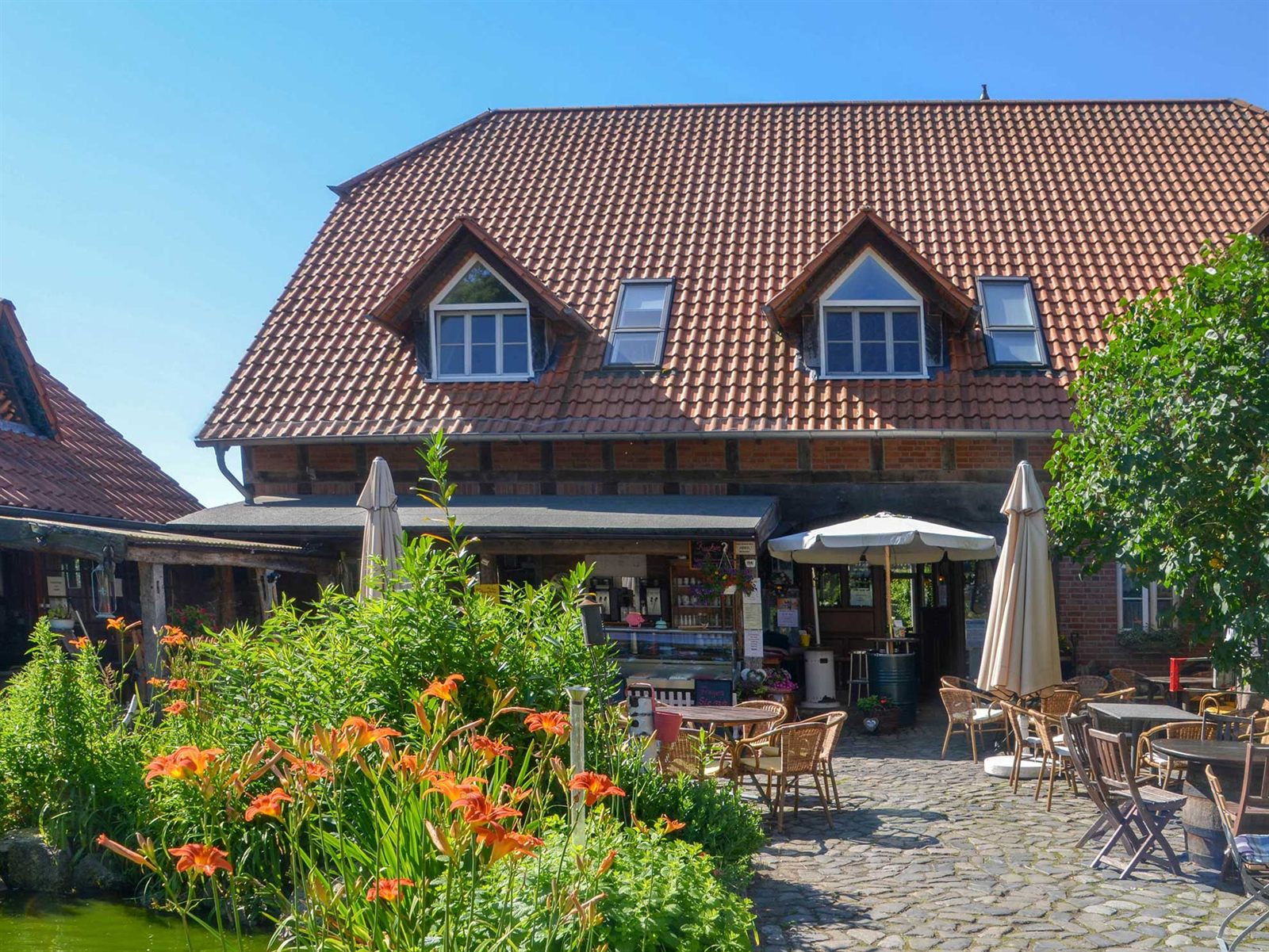 Sonniger Innenhof des Cafés: Fachwerkhaus mit rotem Ziegeldach, Kaffeetheke unter dem Vordach, blühende Lilien und Kopfsteinpflaster