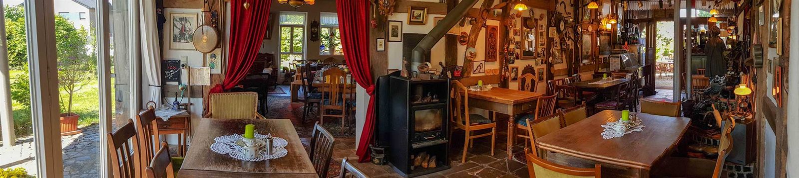 Panoramablick durch die Caféräume: gedeckte Holztische, Kaminofen und Antiquitäten bis unter die Decke