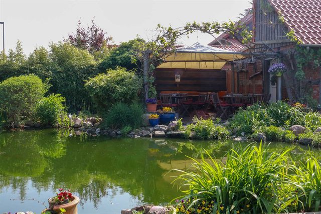 Gartenteich mit Ufersteinen, Gräsern und überdachter Sitzecke unter einer Pergola