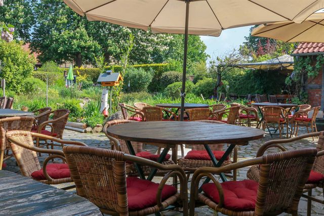 Kaffeegarten mit Korbstühlen, roten Sitzkissen und Sonnenschirmen zwischen blühenden Beeten