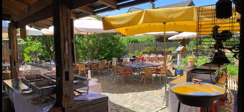 Für eine Feier eingedeckte Tische mit Gläsern im sonnigen Kaffeegarten, davor das Buffet unter dem Balkenvordach, darüber ein gelber Sonnenschirm
